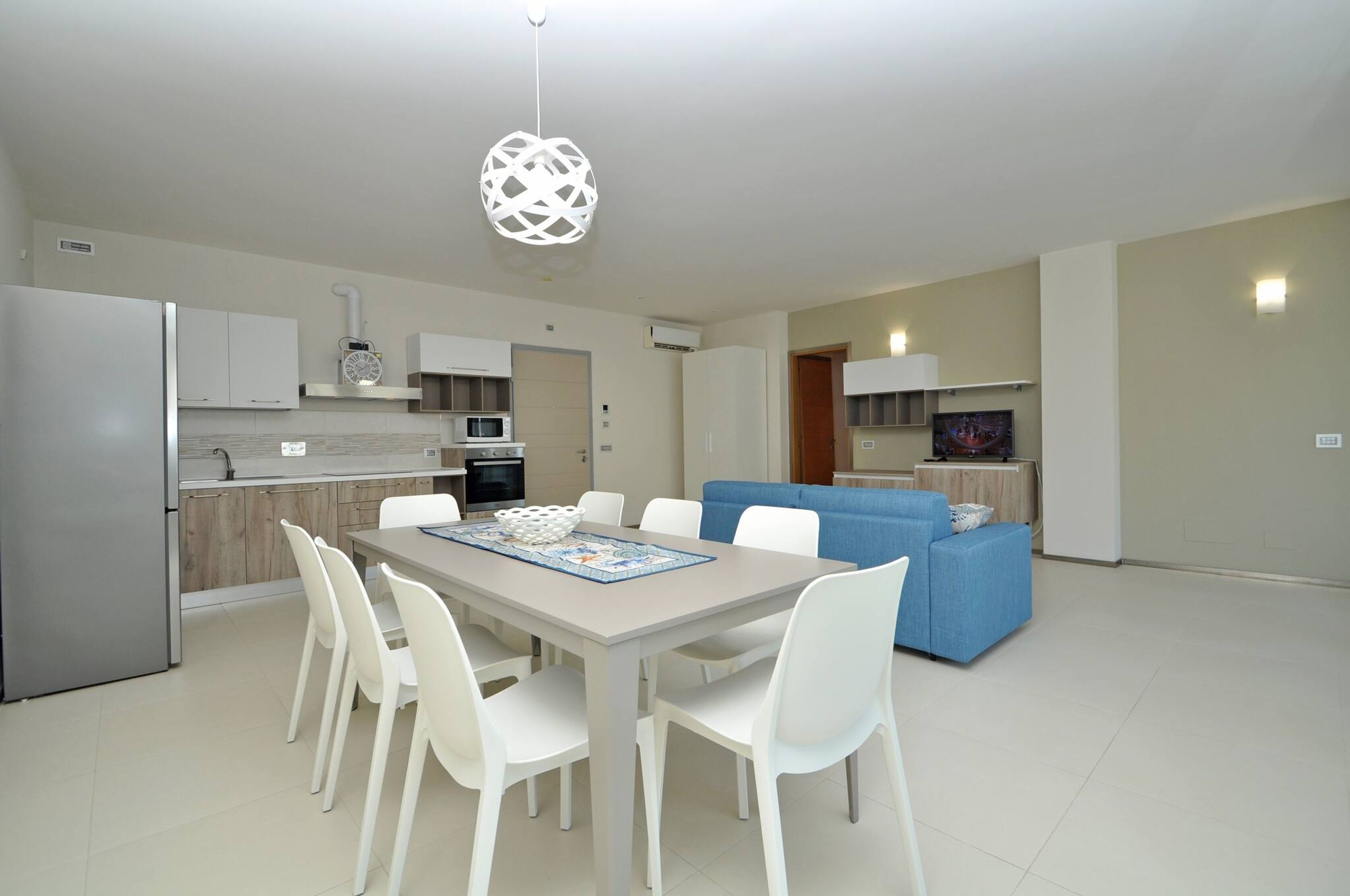 Una cocina moderna con armarios blancos, un refrigerador y un microondas. Hay un comedor con una mesa y sillas, y un sofá azul en la sala.