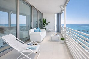 A balcony with a white sofa, a white lounge chair, a small table with a plant, and a view of the ocean.