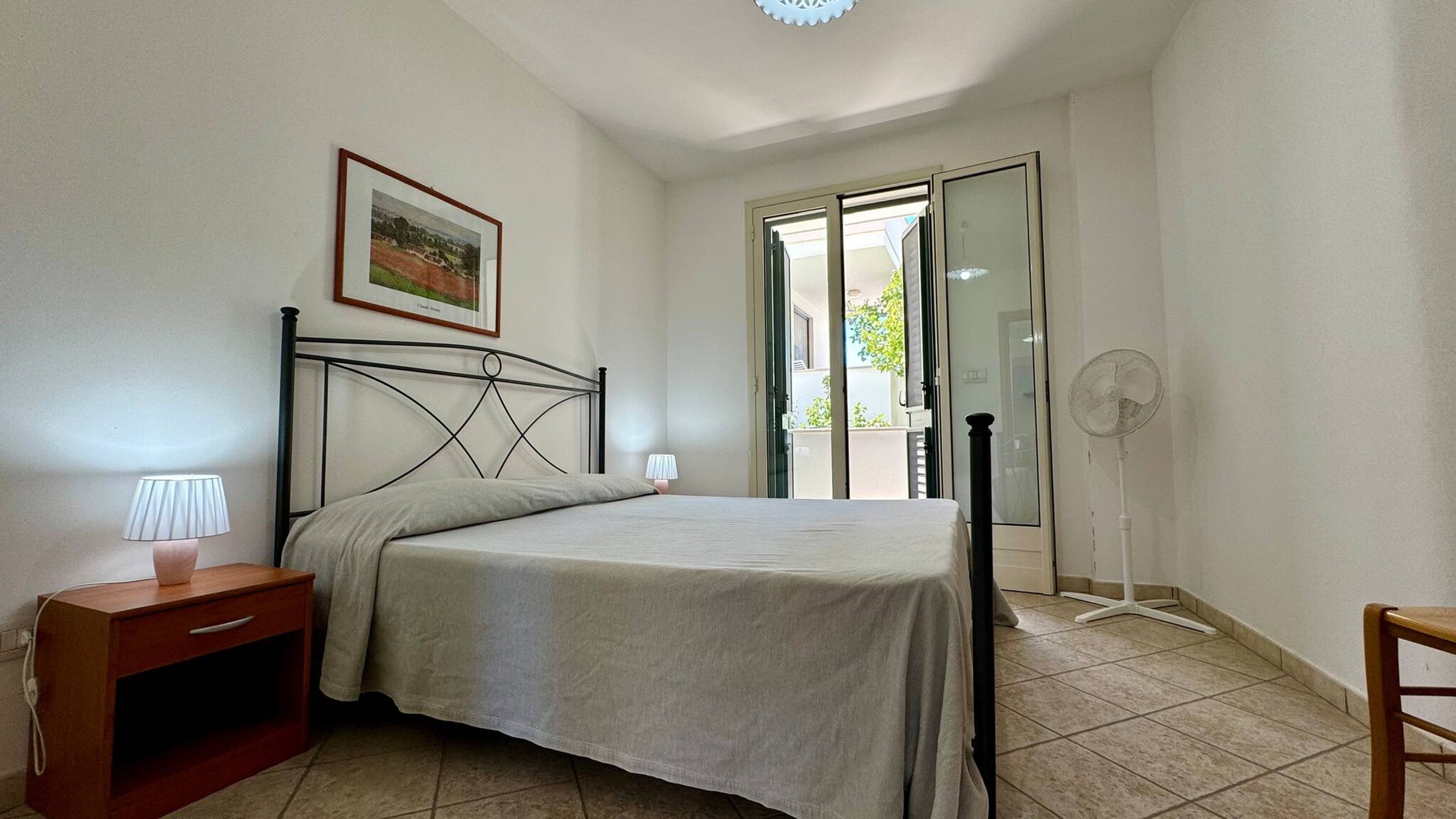 A bedroom with a bed, a nightstand, a lamp, a fan, and a door leading to a balcony.