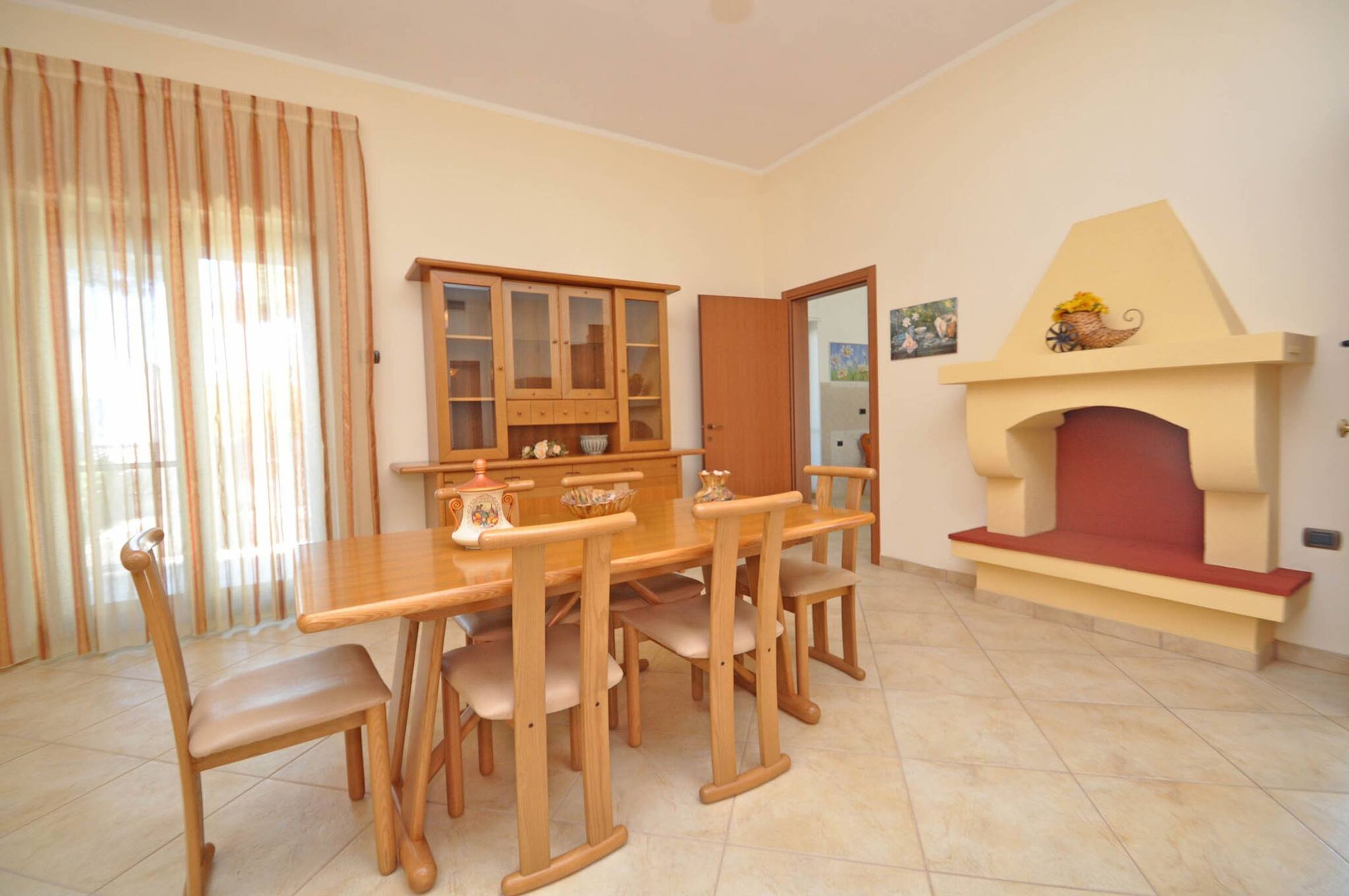A dining area with a wooden table and chairs, a cabinet with glass doors, and a fireplace with a red seat.