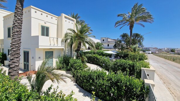 A modern residential building with a white facade, green shutters, and a well-maintained garden with palm trees and shrubs. - Bilocale Arainn 2 - Japigia (Ugento)