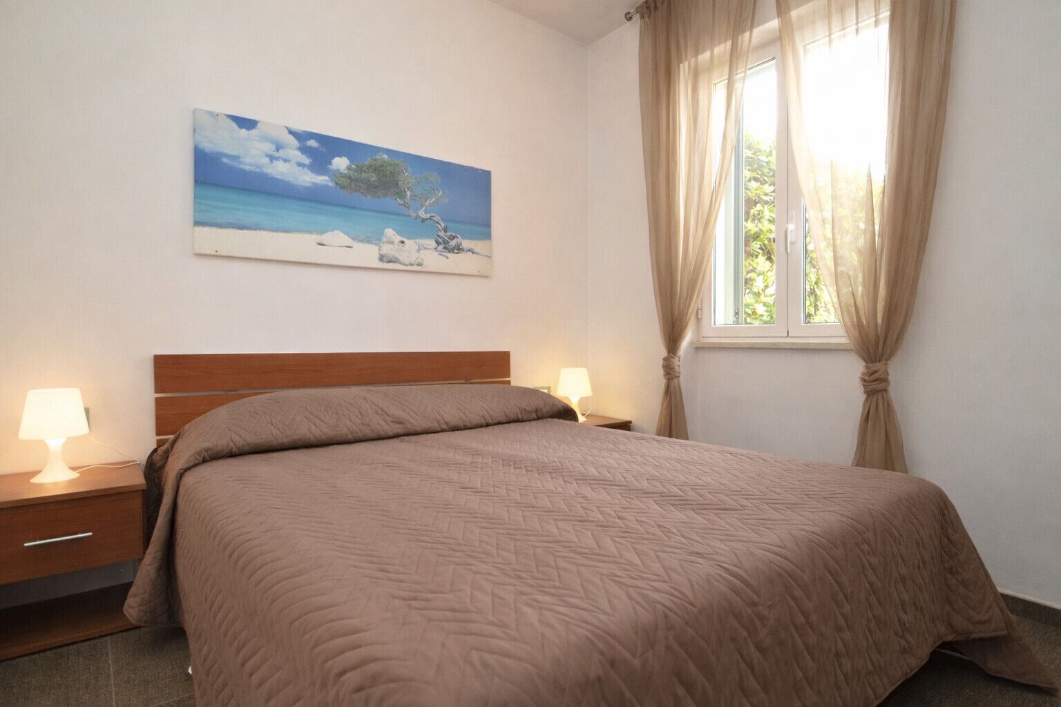 A bedroom with a bed, two bedside tables with lamps, a painting of a beach scene, and a window with curtains.