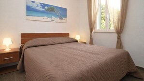 A bedroom with a bed, two bedside tables with lamps, a painting of a beach scene, and a window with curtains. - Bilocale Arainn 2 - Japigia (Ugento)