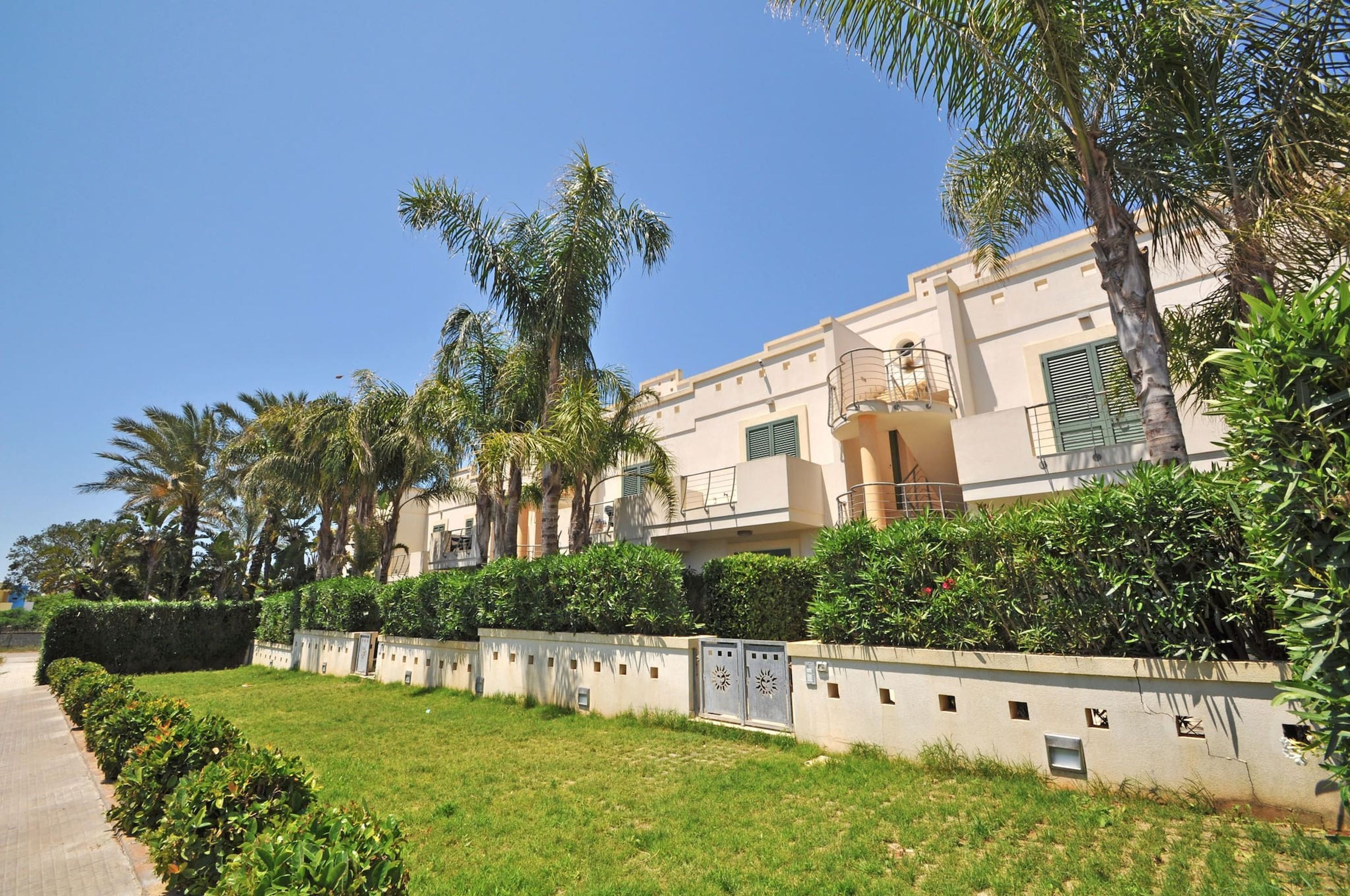 A residential building with multiple balconies, a paved pathway, and lush greenery including palm trees and hedges.