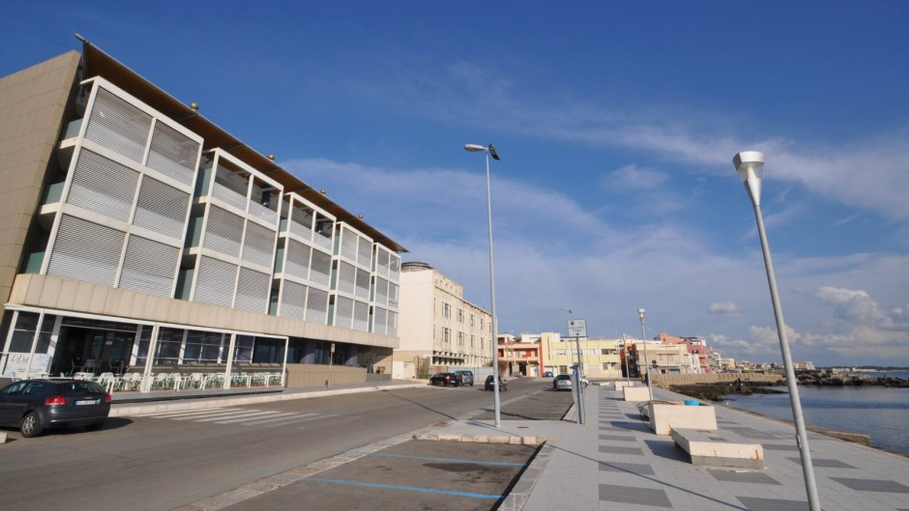 Un bâtiment moderne doté de grandes fenêtres et d’un toit plat, situé près d’une promenade en bord de mer, sous un ciel dégagé.