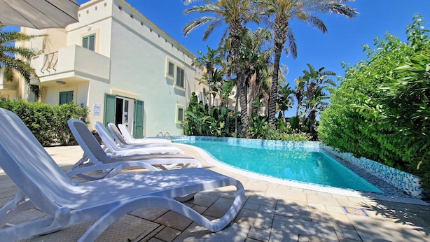 Un espace aménagé au bord de la piscine, avec des chaises longues, une piscine à l’eau d’un bleu limpide et un bâtiment doté de balcons et de palmiers.