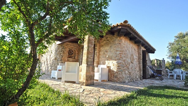 A stone building with a tiled roof, outdoor seating area with white wicker chairs, and a stone pillar. - Trullo Beneficati in Taviano (Taviano)