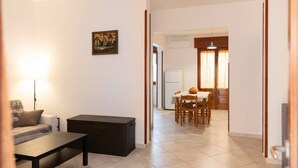 A living room with a sofa, a coffee table, and a painting on the wall. There is a dining area with a table and chairs, and a kitchen visible in the background.