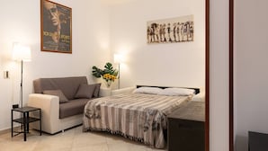 A room with a sofa, a bed with a patterned bedspread, a small table, a floor lamp, and a poster on the wall.