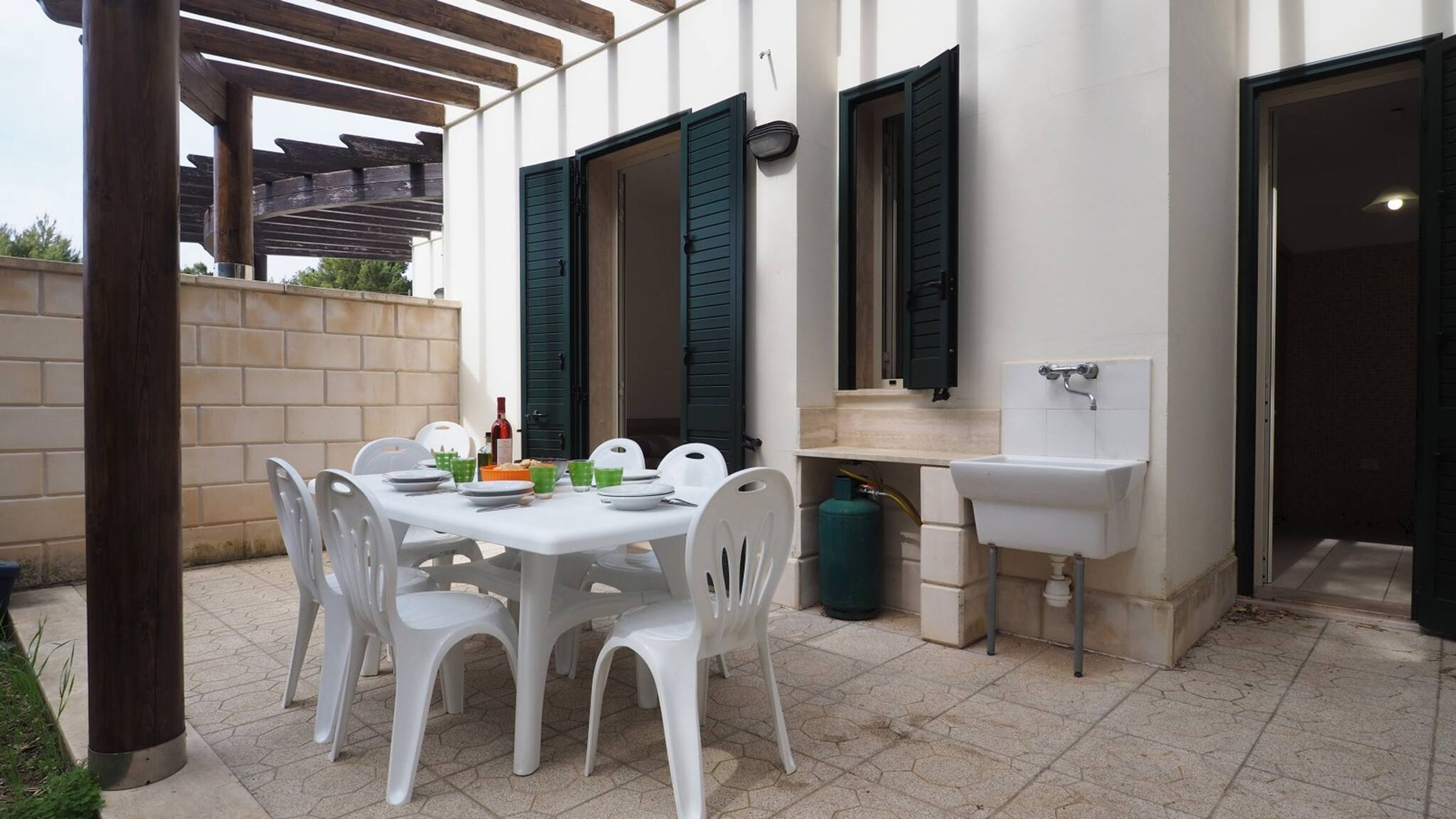 A patio with a table set for four, white plastic chairs, a small sink, and a window with green shutters.