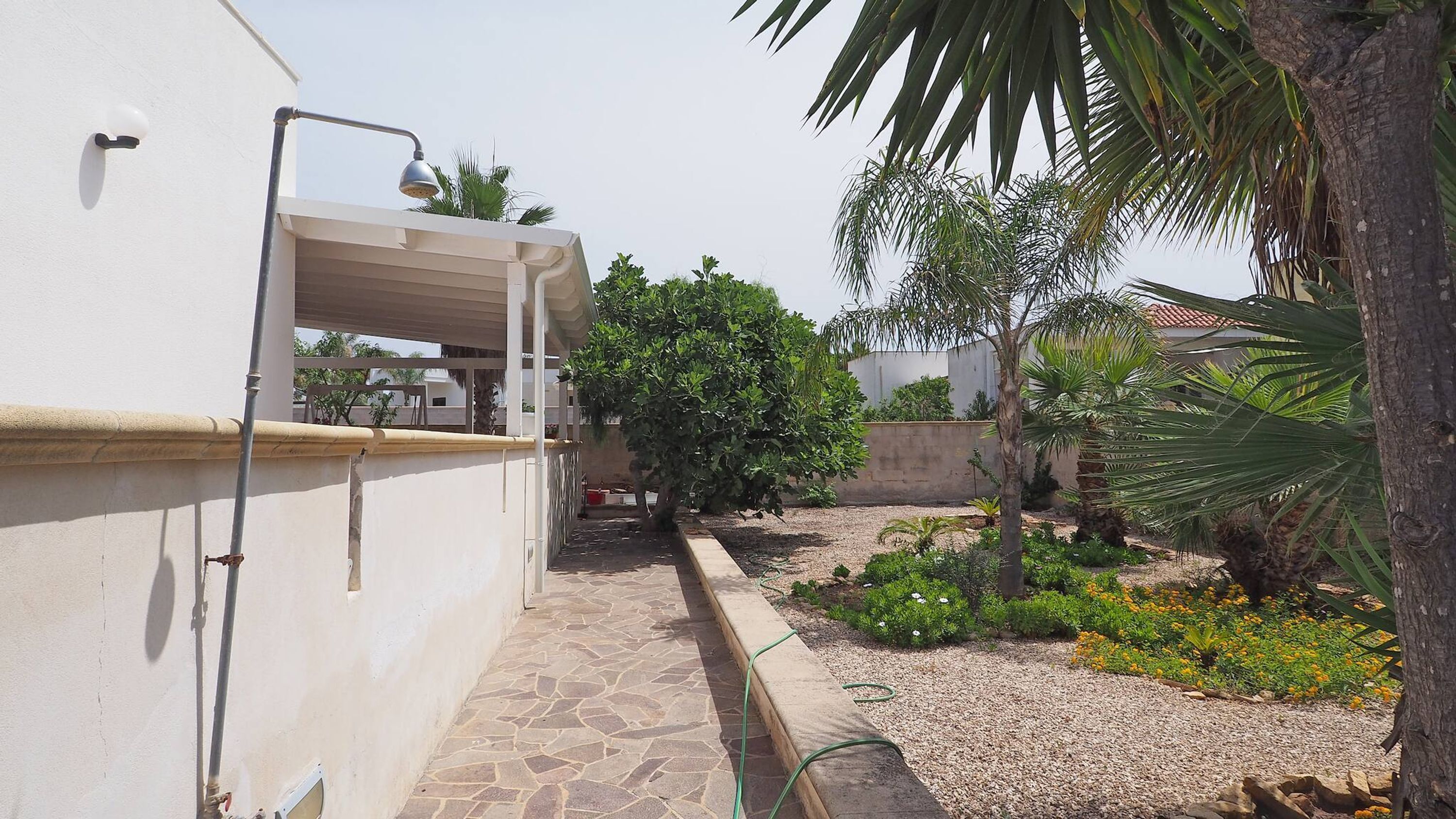 A narrow stone pathway leading to a covered outdoor area with a pergola, surrounded by greenery and a small garden with flowering plants.