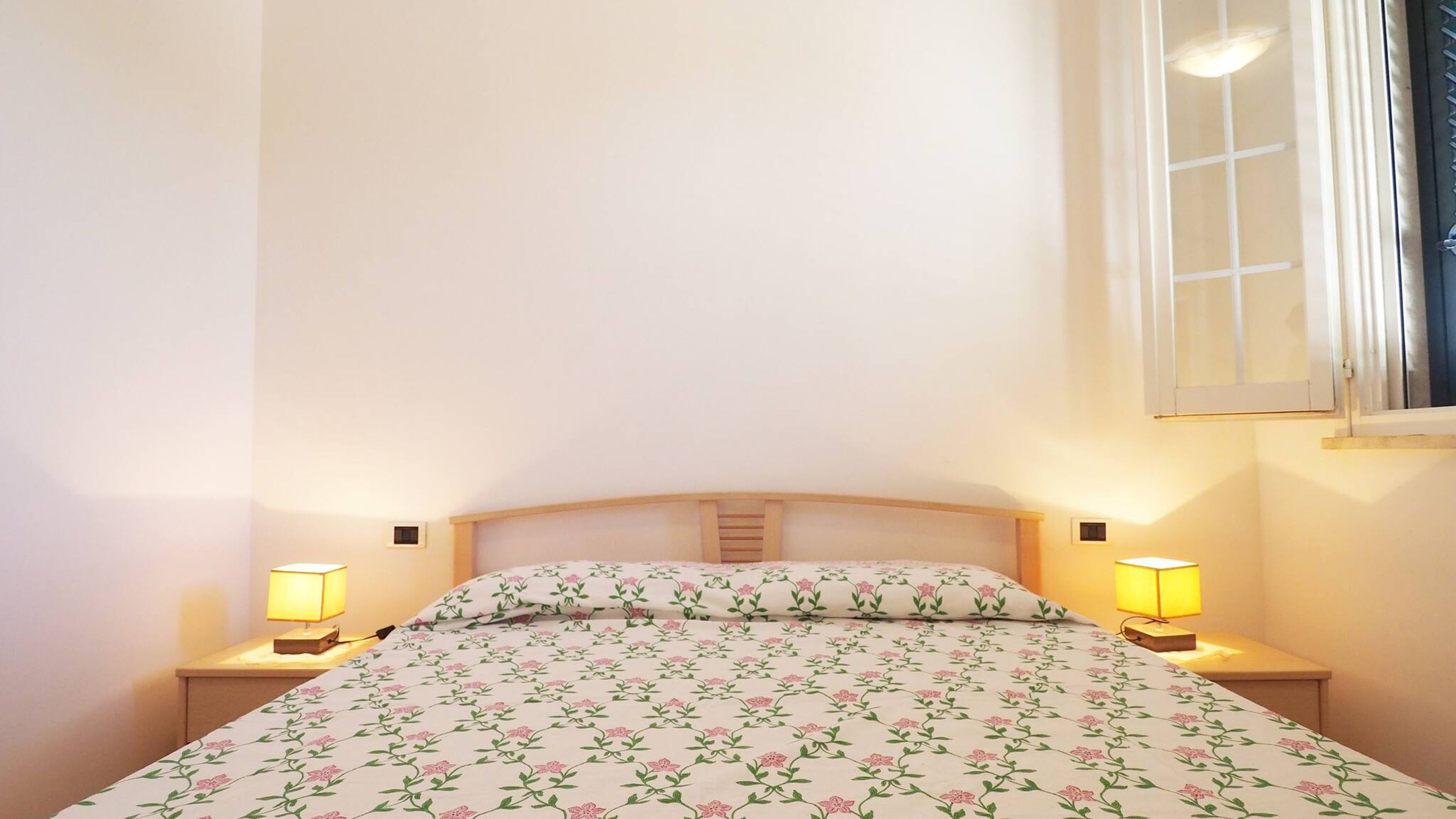 A single bed with a floral bedspread, two bedside lamps, and a window with blinds.