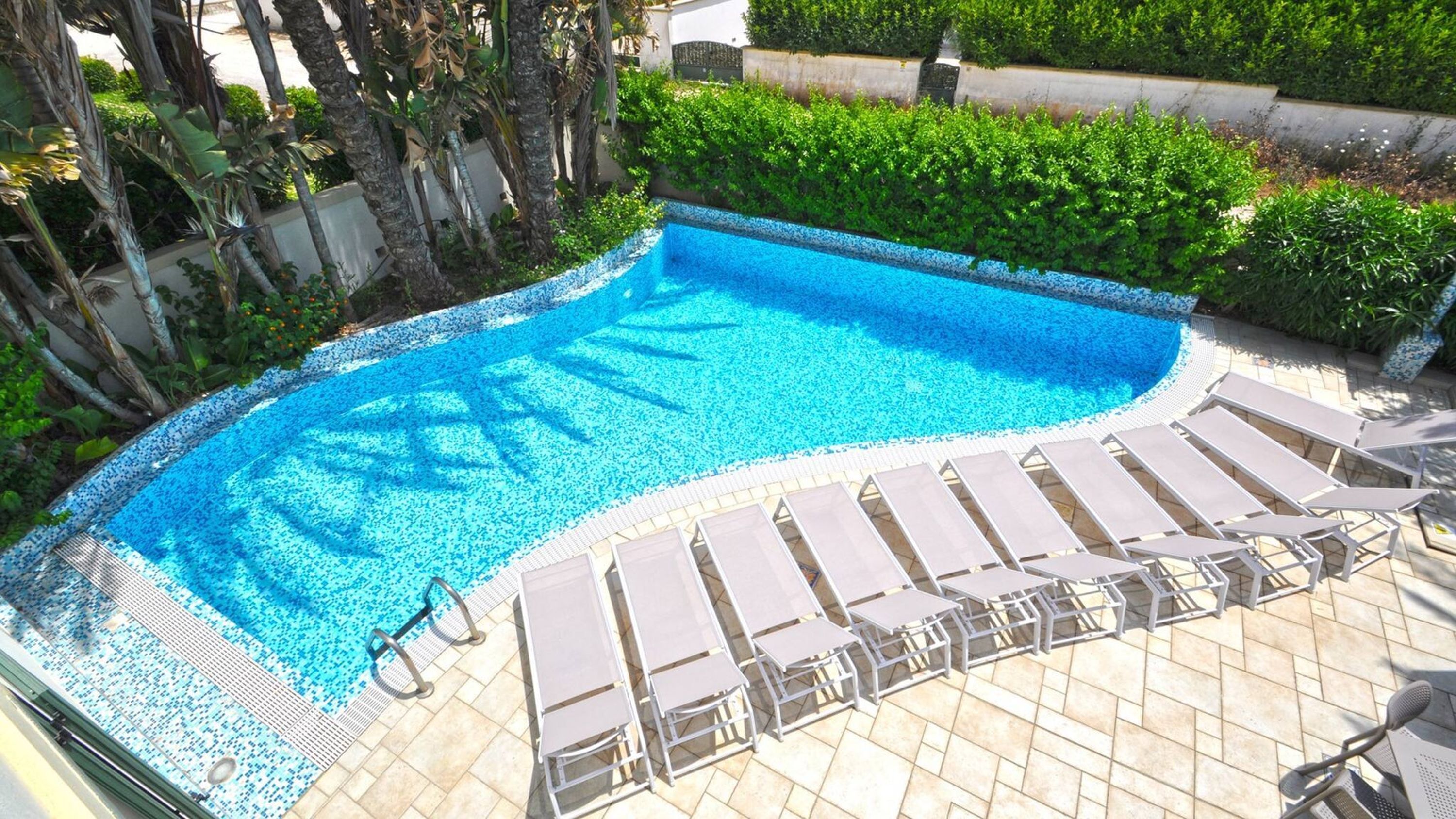 Piscina com deck azulejado, rodeada por vegetação e um muro branco.