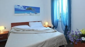 Ein ordentlich bezogenes Bett mit weißen Leinen, ein hölzernes Kopfteil und ein Strandbild darüber. Ein Fenster mit blauen Vorhängen, eine Nachttischlampe und ein Blumenstrauß auf dem Nachttisch sind ebenfalls zu sehen.