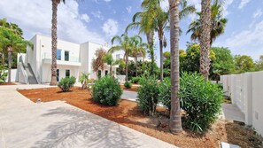 Un bâtiment moderne à la façade blanche, doté d’un espace extérieur couvert et d’un jardin aménagé avec des palmiers et des arbustes.
