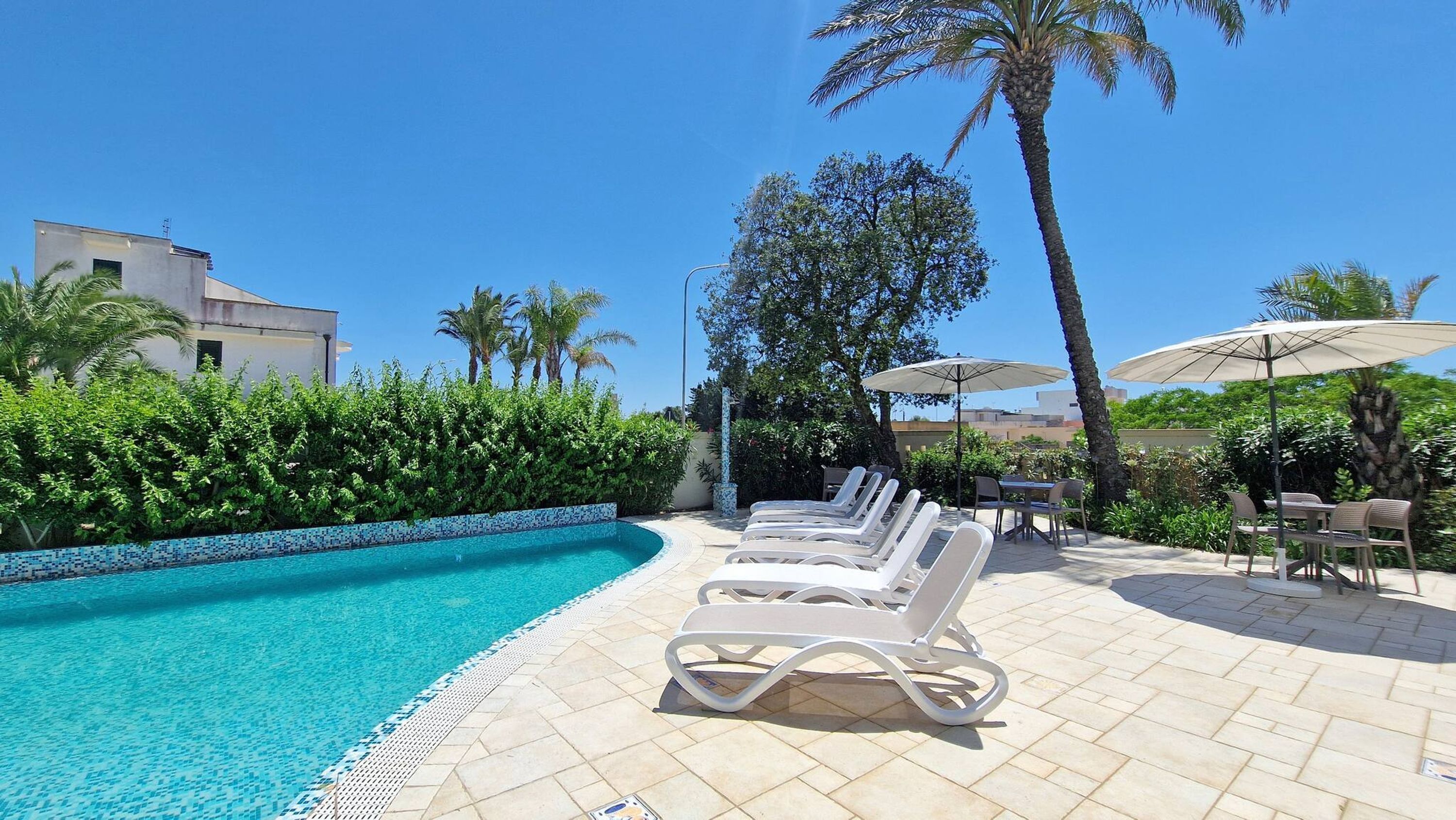 A pool area with lounge chairs and umbrellas, surrounded by palm trees and a tiled pool edge.