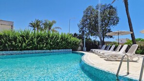 A swimming pool with clear blue water, surrounded by a tiled edge and a row of white lounge chairs with umbrellas.