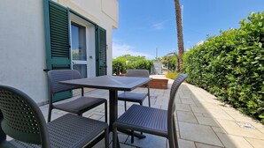 Eine Terrassenausstattung mit Tisch und Stühlen, eine Tür mit grünen Fensterläden und ein Heckenstreifen.