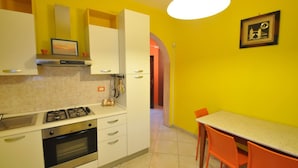 A kitchen with white cabinets, a gas stove, and an oven. There is a dining table with orange chairs and a yellow wall. - Villa for 6 People 2 Bedrooms a few Meters From the sea (Ugento)