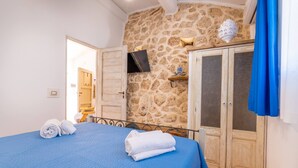 A bedroom with a stone wall, a bed with blue bedding, a television mounted on the wall, and a wardrobe. - Trullo Mmammineddhu in Torre San Giovanni (Ugento)