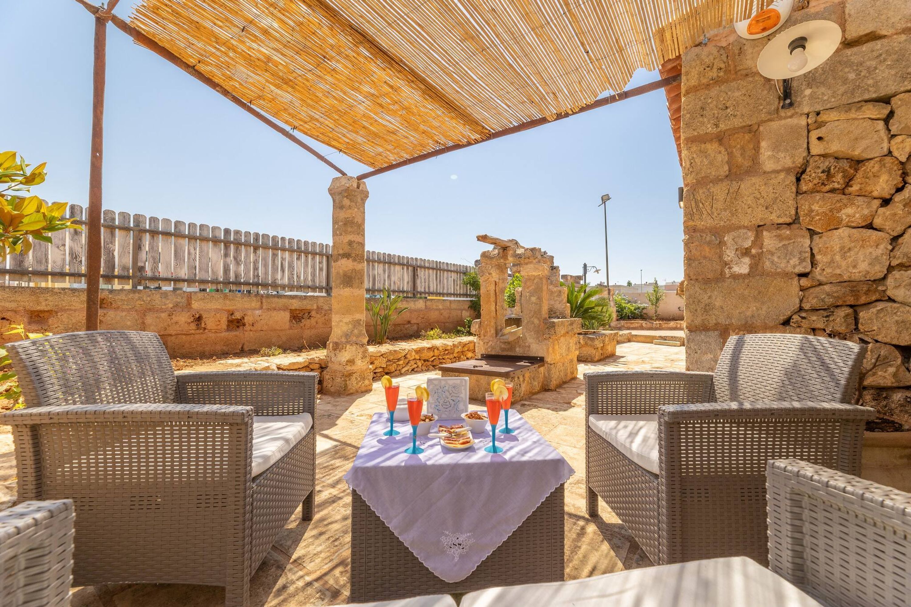 An outdoor patio with wicker furniture, a table set with drinks, and a stone wall in the background.