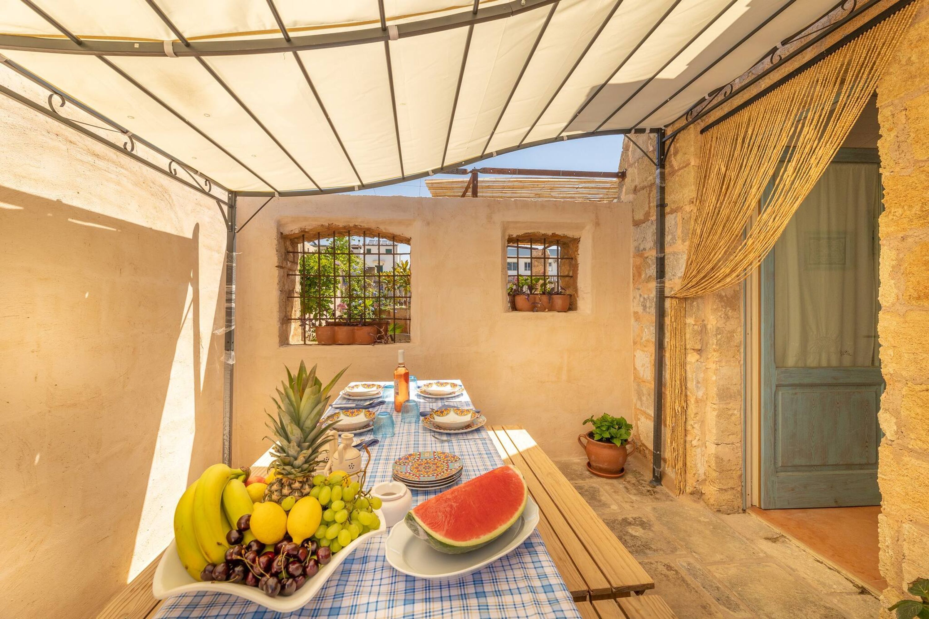 A covered outdoor dining area with a table set for two, featuring a variety of fruits and a bottle of water.