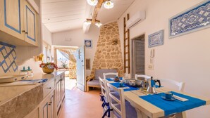 A cozy kitchen with a stone wall, wooden cabinets, and a dining area with blue tablecloths and white chairs. - Trullo Mmammineddhu in Torre San Giovanni (Ugento)