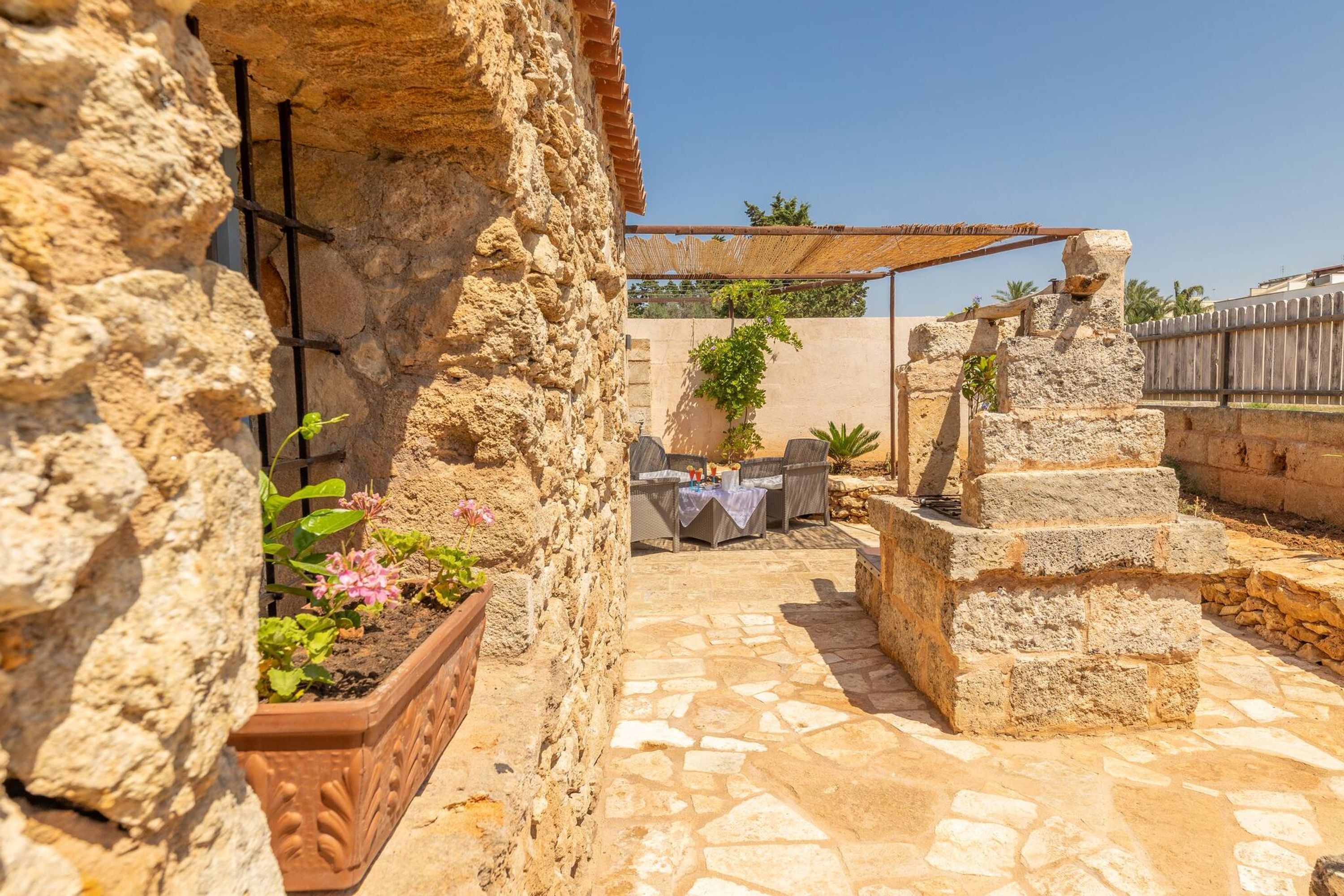 A stone courtyard with a table and chairs under a pergola, surrounded by stone walls and potted plants.