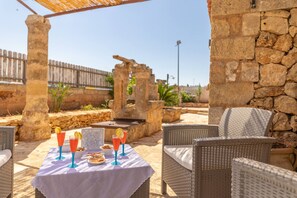 Un patio al aire libre con mobiliario de mimbre, una mesa con bebidas y aperitivos, y paredes de piedra.