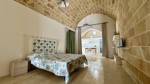 A bedroom with a bed, a nightstand, a TV on a shelf, and a view of a kitchen through an archway. - App al Castello 1 by Salentoaffitti (Alliste)