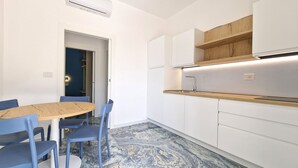 A modern kitchen with white cabinets, a wooden countertop, and a built-in oven. There is a dining area with a round table and blue chairs. - Suite Fontanelle Luxury 8 (Ugento)