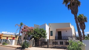 A modern two-story house with a balcony, surrounded by a black metal fence, palm trees, and a clear blue sky. - Villetta Torre Suda 1 (Racale)