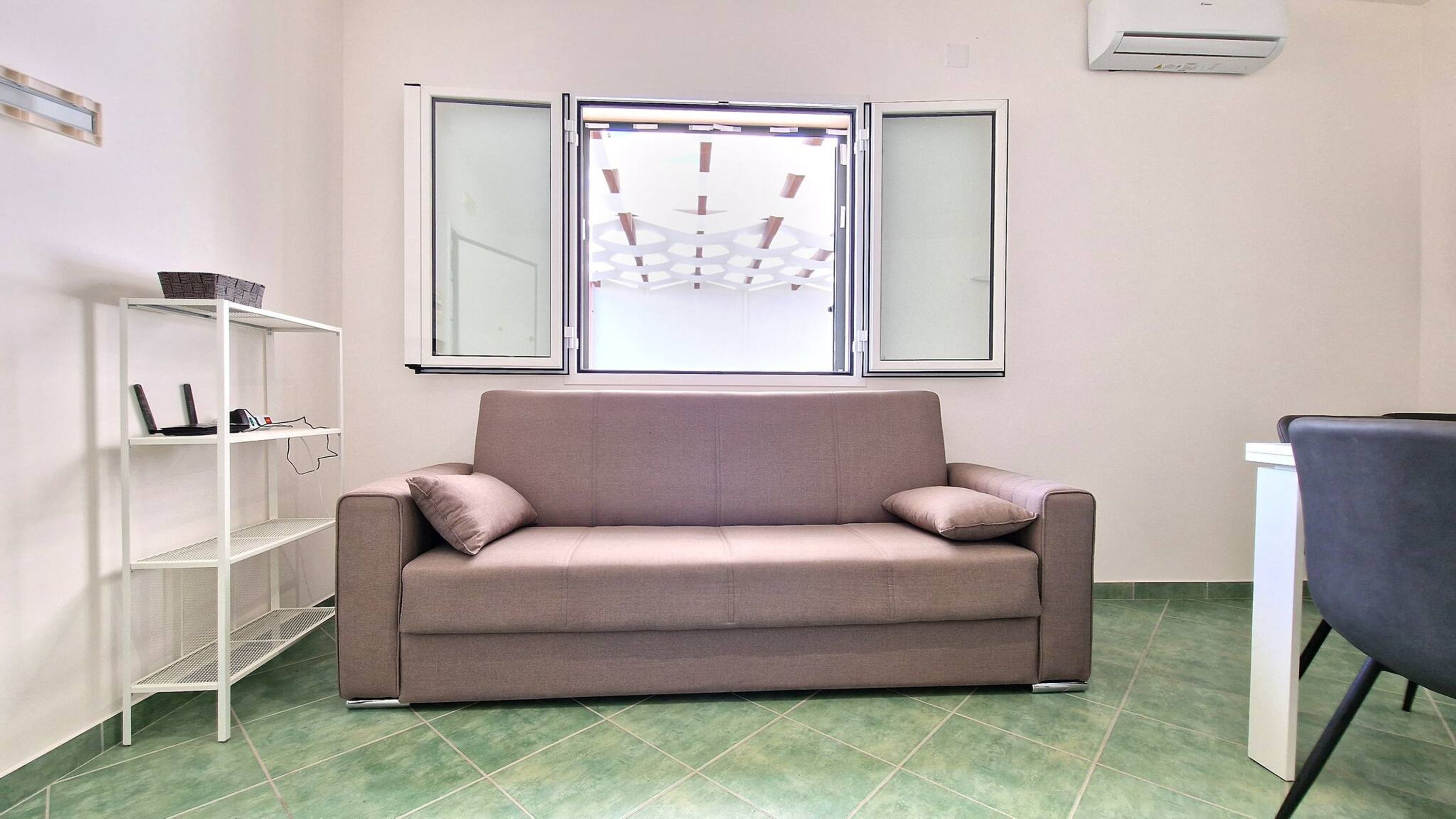 A room with a beige sofa, a white shelving unit with electronic devices, and a window with a geometric view.