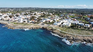An aerial view of a coastal residential area with houses along the shoreline, a road, and a rocky breakwater. - Villetta Charme 2 in Capilungo 5 Guests by the sea (Alliste)