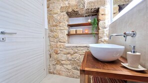 A bathroom with a stone wall, a wooden vanity, a white sink, and a shelf with candles and a plant. - Tenuta Alle Macchie in Racale (Racale)