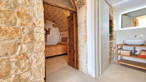 A stone-walled room with a bed, a wooden door, and a bathroom with a sink and mirror. - Tenuta Alle Macchie in Racale (Racale)