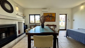 A dining area with a long table, chairs, a fireplace, a painting, and a view through the window. - Villetta Certosina 1 (Alliste)