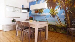 Ein Essbereich mit einem weißen Tisch und Stühlen, einem an der Wand montierten Fernseher und einem Wandbild mit Strandmotiv.