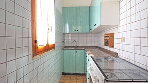 Egy kis méretű konyha fehér csempézett falakkal, függönnyel ellátott ablakkal, türkiz színű szekrényekkel, mosogatóval és tűzhellyel.