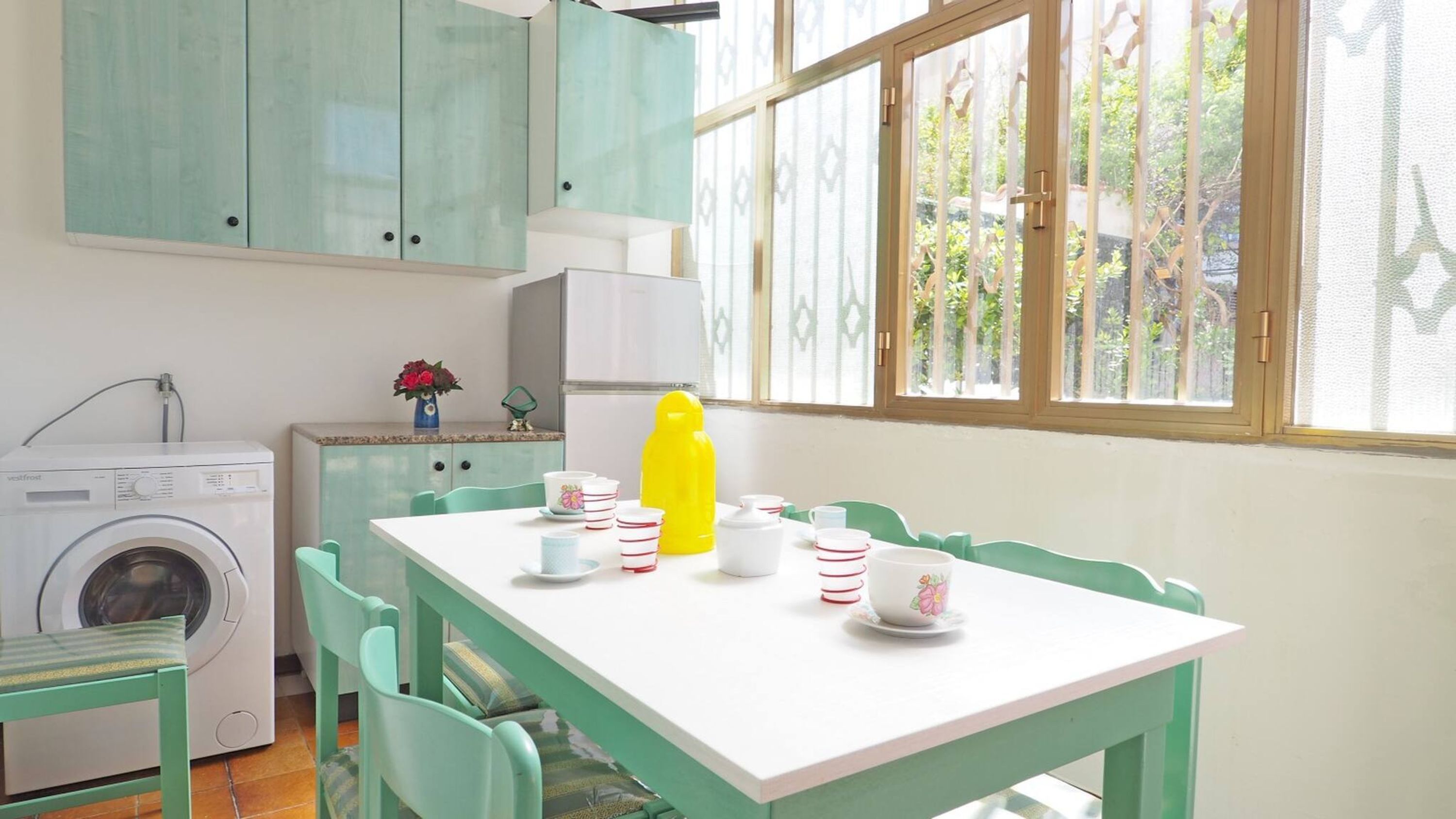 Une cuisine compacte équipée d’un lave-linge, d’une table à manger avec des chaises et d’une fenêtre donnant sur un espace verdoyant.