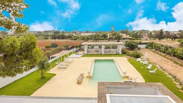 Un área de piscina con sillas de descanso y una pérgola, rodeada de césped cuidado y árboles.