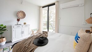 A modern bedroom with a large bed, a white wardrobe, a small table with decorative items, and a view of the outdoors.