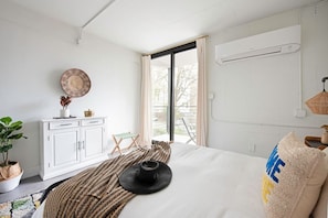 Une chambre à coucher moderne avec un grand lit, une armoire blanche, une petite table agrémentée d’objets décoratifs et une vue sur l’extérieur.