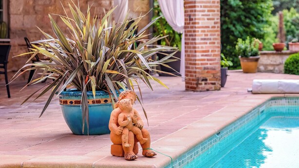 Un espace aménagé au bord de la piscine, avec une statue, une plante en pot et un bâtiment en briques en arrière-plan.
