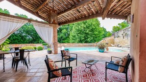 Un espace extérieur aménagé, abrité et doté de chaises et de tables, une piscine et une végétation luxuriante en arrière-plan.