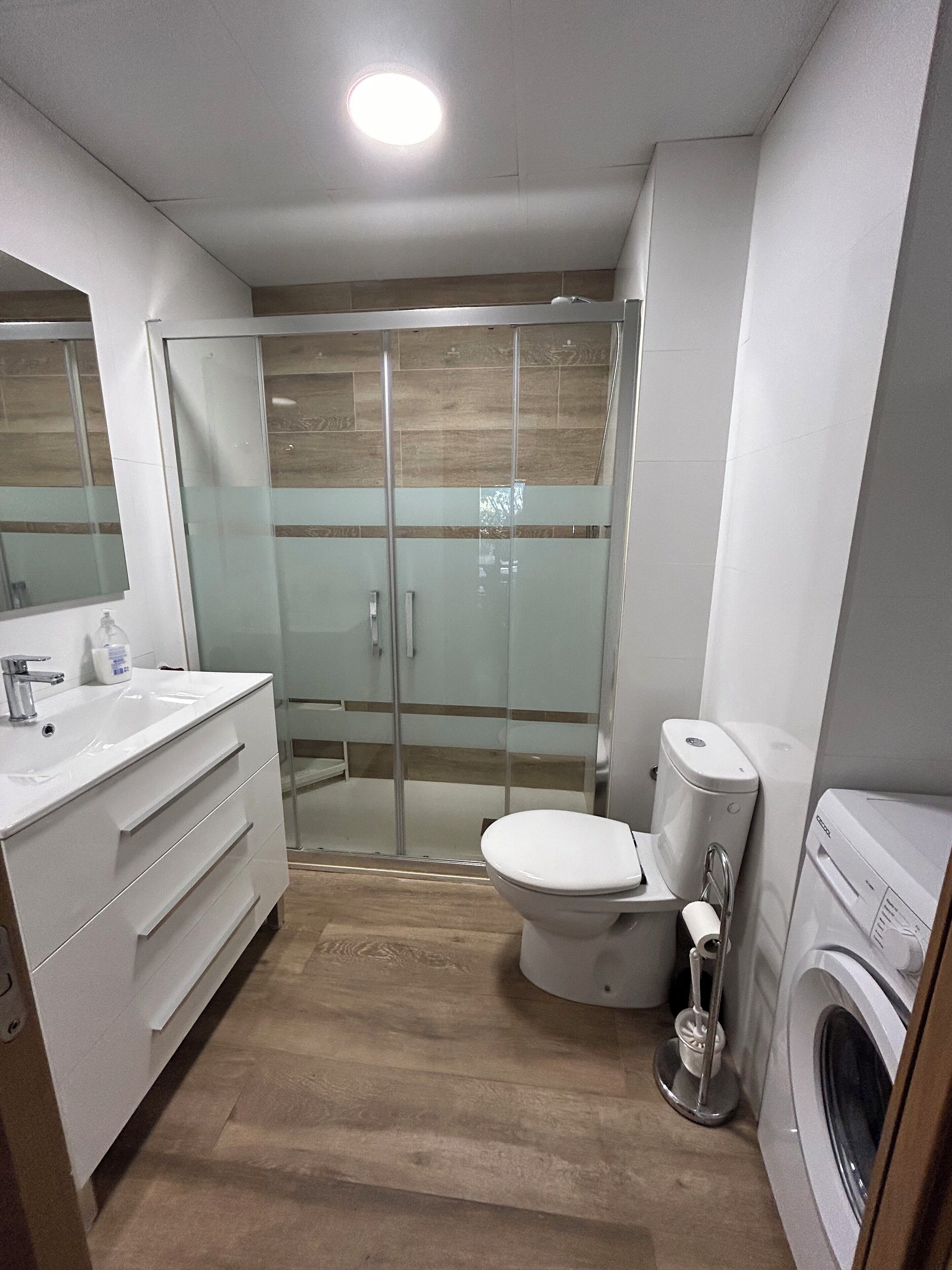 A modern bathroom with a glass shower enclosure, a toilet, a sink, and a washing machine.