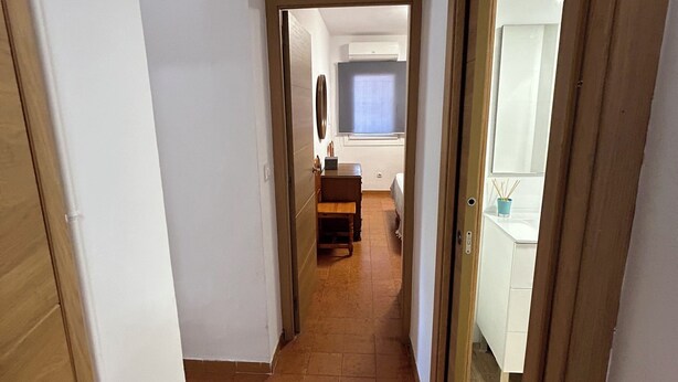 Un couloir étroit, dont le sol est recouvert de carreaux, mène à une pièce où se trouvent un lit et une fenêtre.