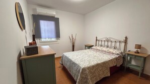 Makuuhuoneessa on sänky, lipasto, yöpöytä, lamppu, ikkuna, jossa on verho, ja ilmastointilaite.