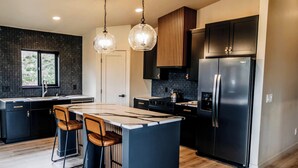 Cocina moderna con armarios oscuros, una isla central con taburetes y un refrigerador de acero inoxidable, además de iluminación colgante.