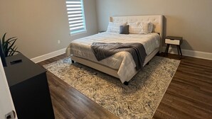 Une chambre à coucher avec un lit, un ventilateur de plafond, une table de chevet, une fenêtre et une commode de couleur foncée.